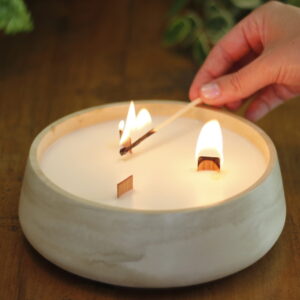 Vela artesanal branca com três pavios de madeira, colocada em um pote redondo de Jesmonite de cor clara. Uma mão está acendendo um dos pavios com um fósforo. A cena se passa sobre uma superfície de madeira, cercada por folhagens verdes, criando uma atmosfera natural e acolhedora.
