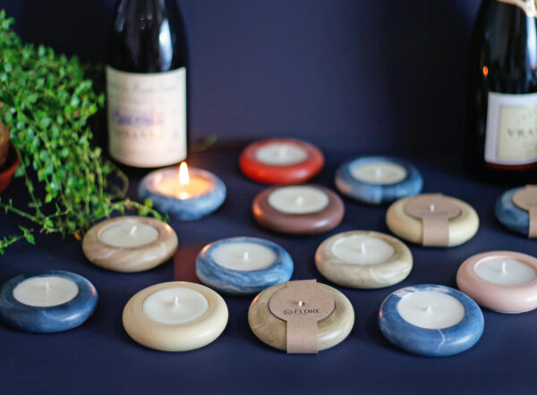 Velas Íris Élore em diversas cores e bandejas, com garrafas e folhagens ao fundo.